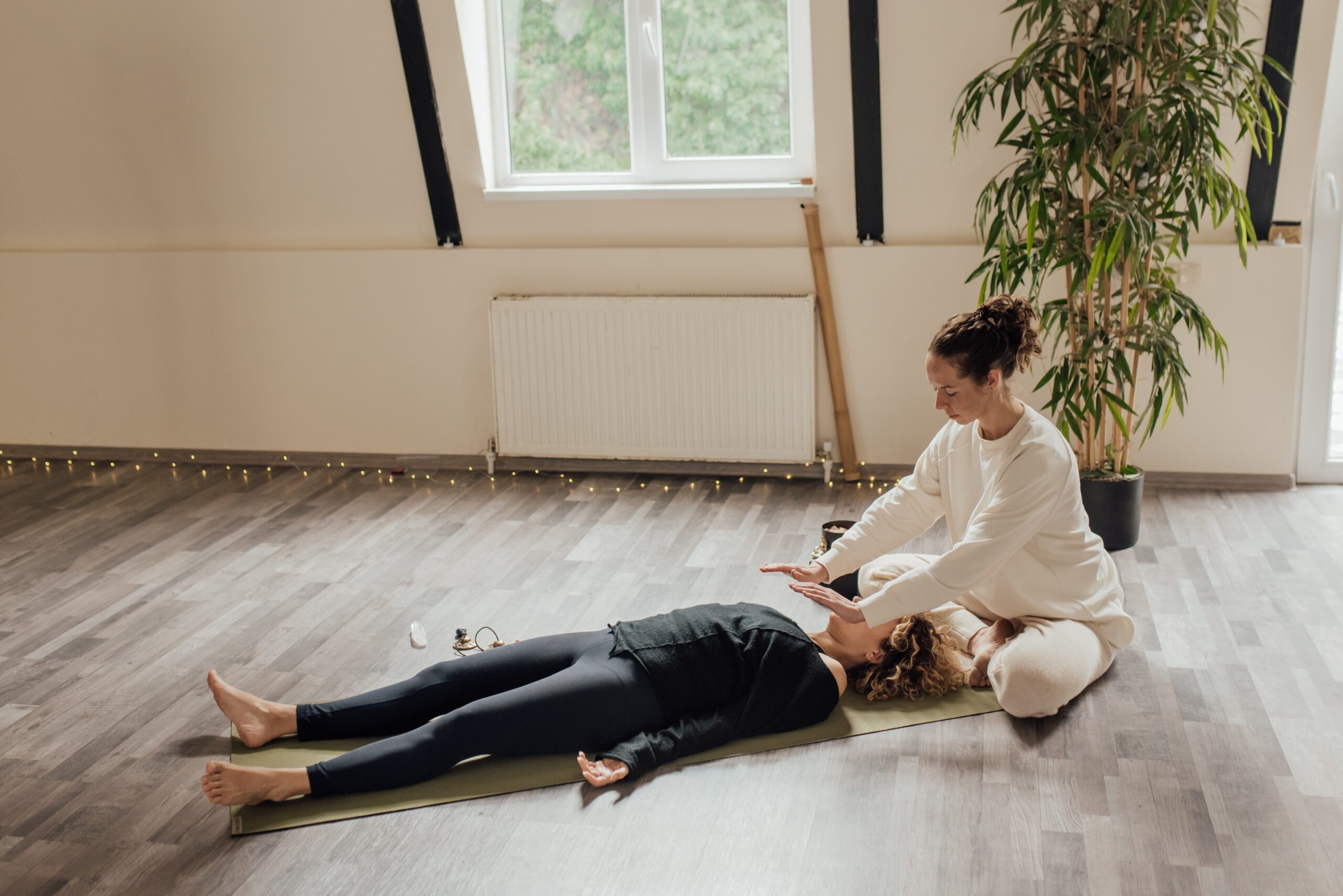 A person lying on a yoga mat in a white room with wooden floors and a plant having an energy healing alternative therapy